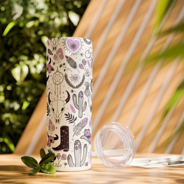 Boho Western Vibes Skinny Tumbler in an outdoor setting, with a rustic background. The tumbler features a bold design of bull skulls, black cowgirl boots, dreamcatchers, and horseshoes in black, white, and lavender, perfect for adventurous spirits.