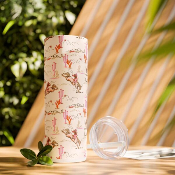 Boho-inspired Cowgirl Skinny Tumbler in an outdoor setting, featuring a bucking horse, pink cacti, and mountain designs in vibrant colors, with a clear lid and matching straw, perfect for on-the-go hydration.